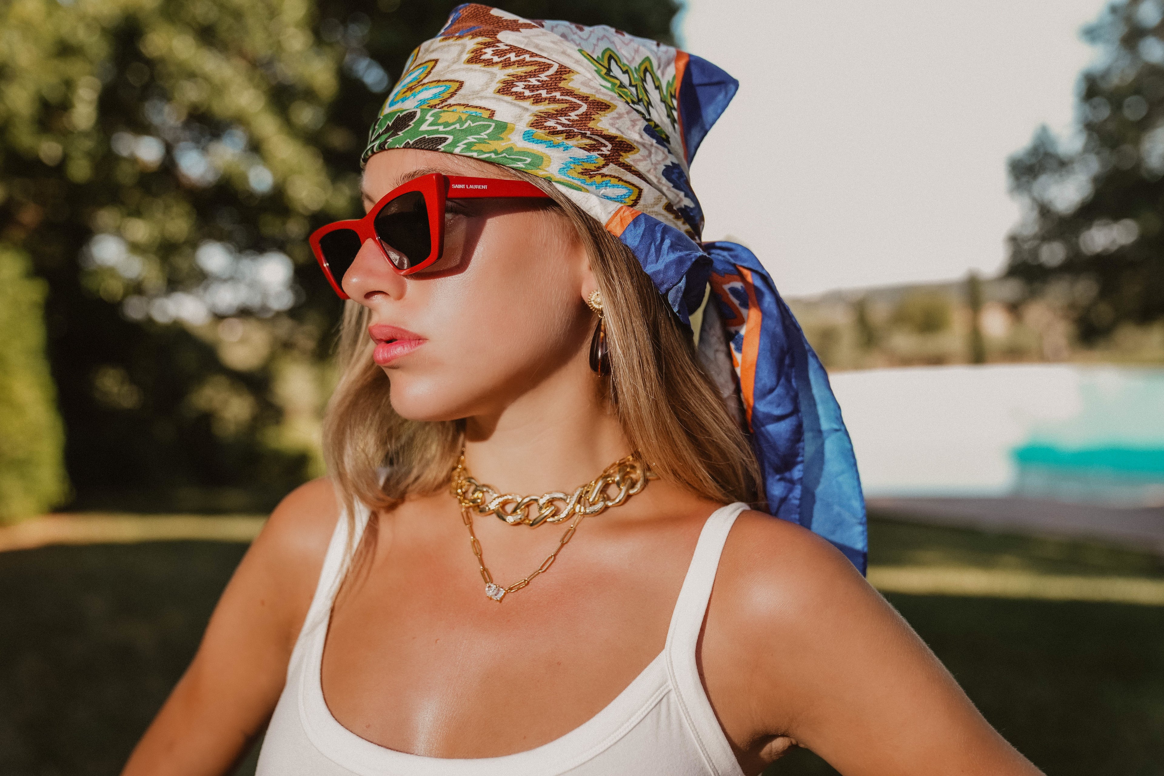 Woman wearing a colorful headscarf, red sunglasses, and gold jewelry outdoors.