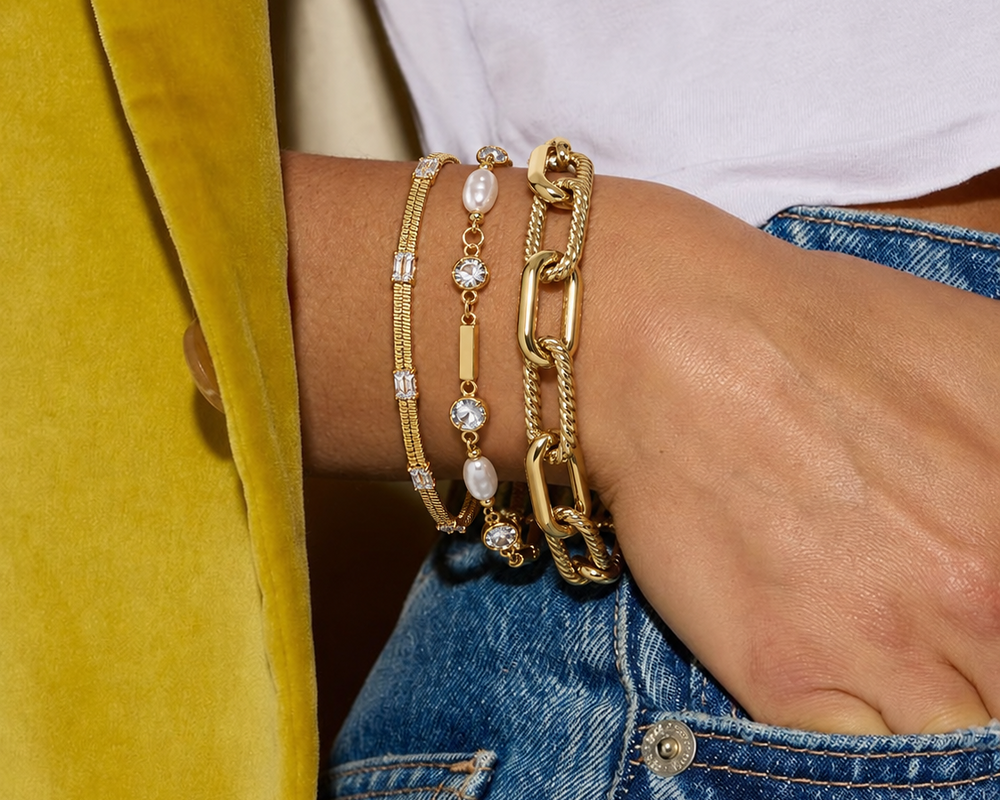 A close-up of a person's wrist wearing a stack of gold bracelets, including a thick chain link bracelet, a rope bracelet, and a delicate beaded bracelet with clear stones, styled with a white top and blue jeans.