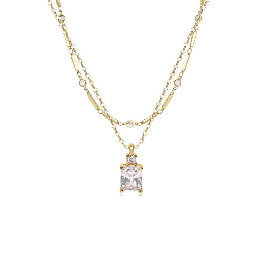 A gold double-strand necklace featuring a rectangular clear gemstone pendant with a bezel setting, accented by small round stones along the chain, shown against a plain white background.